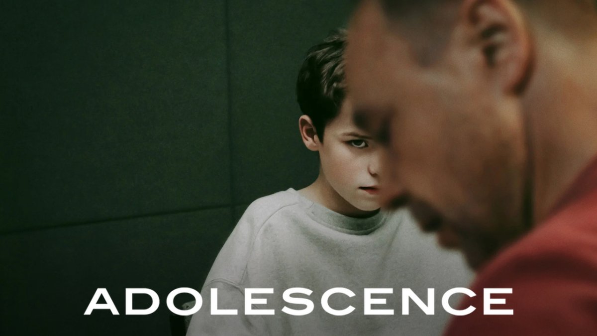《Adolescence》劇集畫面：穿淺色上衣的少年站在前景模糊的紅衣男子身後