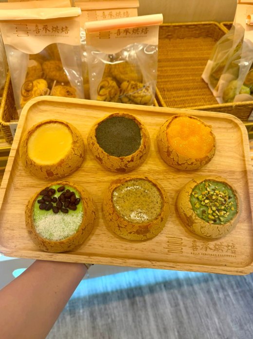 木盤上擺放六個不同餡料與配料的小圓麵包，背景可見包裝烘焙產品